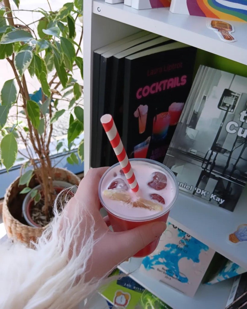 A cocktail in someone's hand. In the background, a shelf of books and a potted plant.
