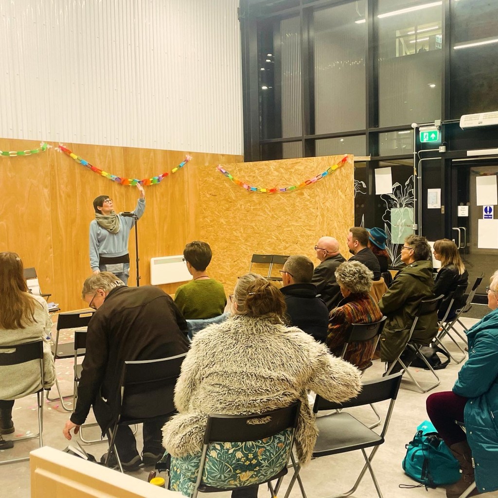 Seated audience watch a poetry performance by Jemima Hughes.