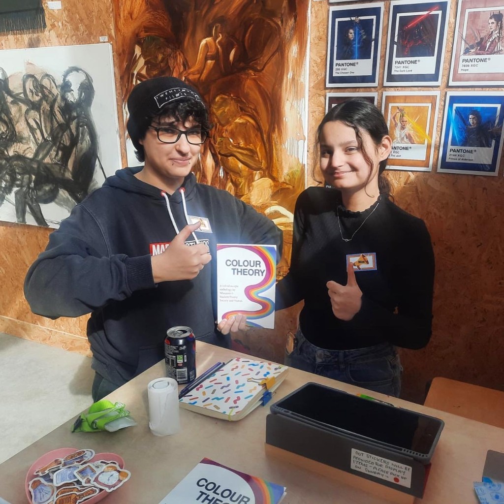 Two volunteers stand in front of a wall of artwork and smile with thumbs up.
